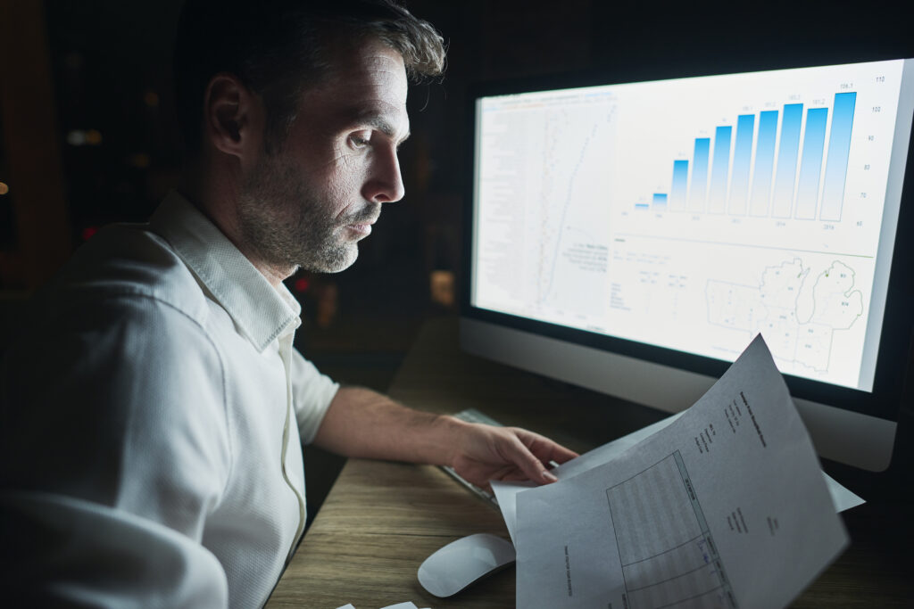 Hombre con camisa blanca, frente a ordenador y con papeles en la mano analizando datos en tiempo real. la clave esencial de ayuda para emprendedores para alcanzar el éxito.